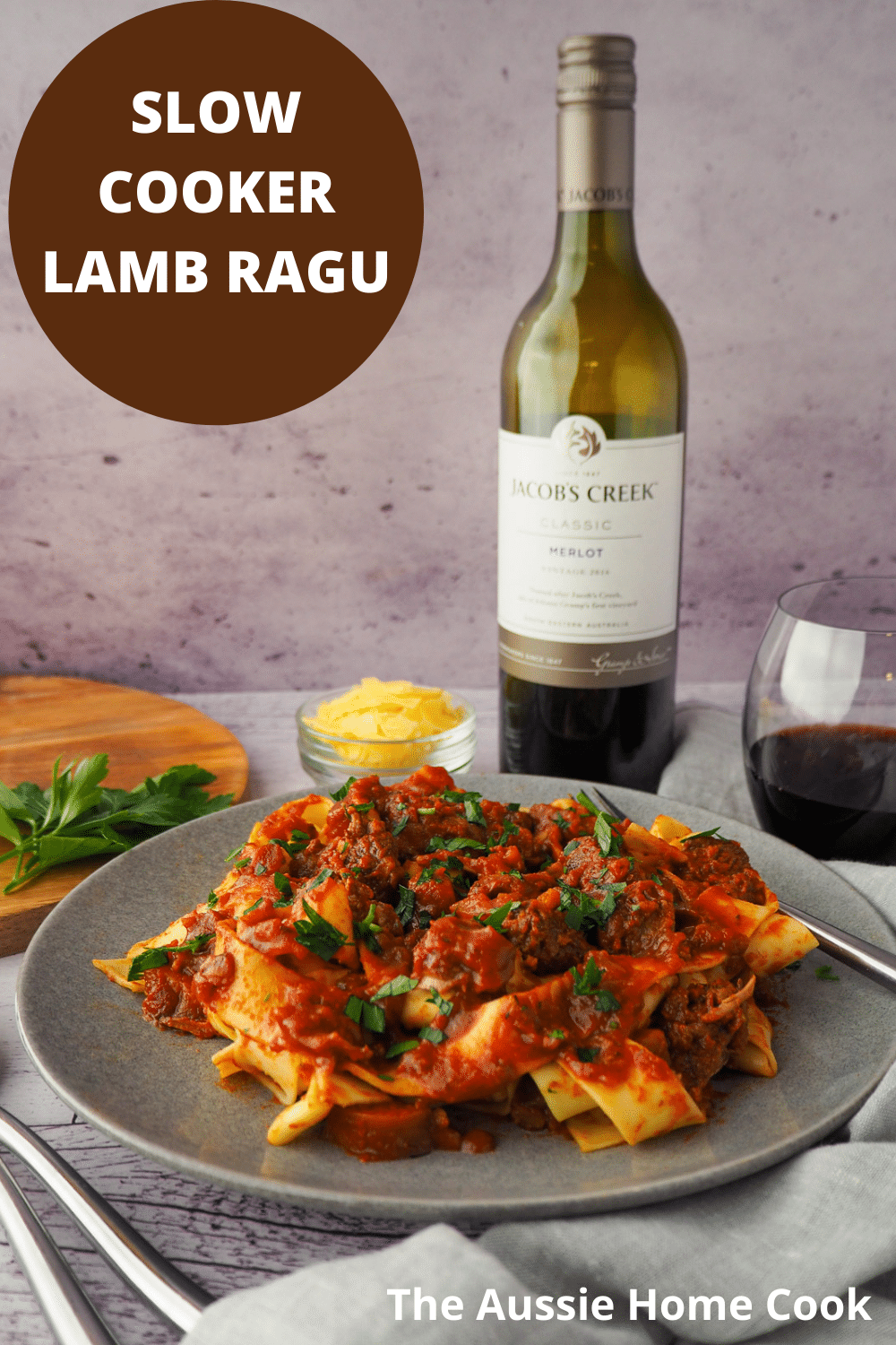 Slow cooker lamb ragu 29 Plate of slow cooker lamb ragu, garnished with chopped parsley, surrounded by serving cutlery, glass of red wine, bowl of parmesan cheese and chopping board with parsley on it and text overlay, slow cooker lamb ragu, The Aussie Home Cook.
