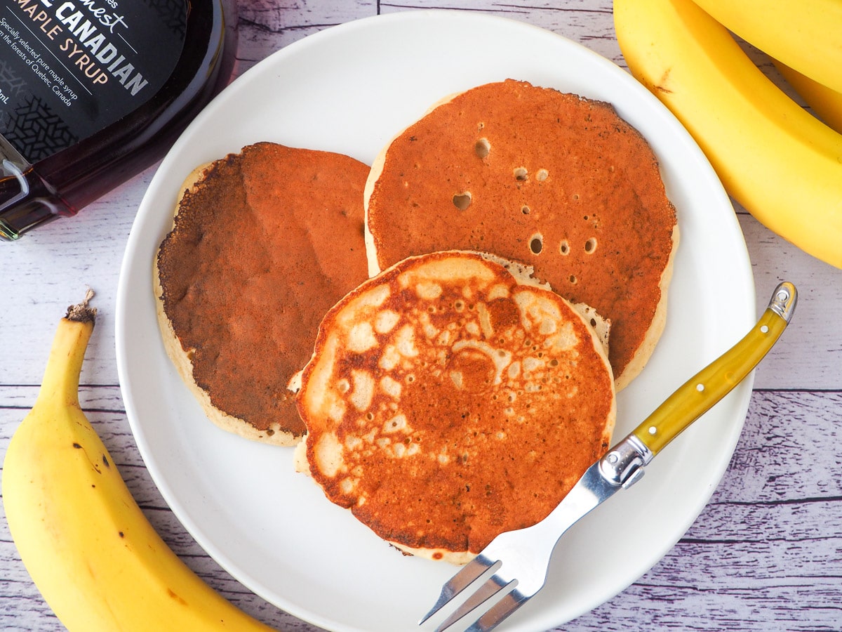 Banana pikelets 14 Top down view of three banana pikelets, surrounded by fresh whole bananas and a bottle of maple syrup.