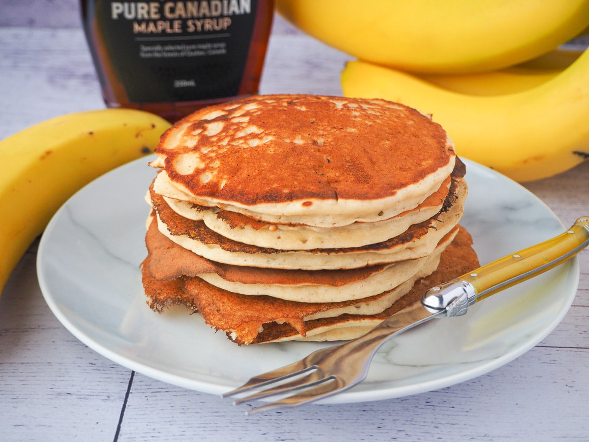 Banana pikelets 1 Stack of banana pikelets on a plate, with a vintage spoon on the side, surrounded by fresh whole bananas and bottle of maple syrup in the background.