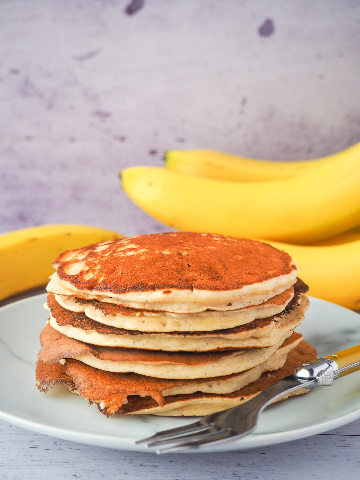 Banana pikelets 18 Stack of banana pikelets on a plate, with a vintage spoon on the side, surrounded by fresh whole bananas.