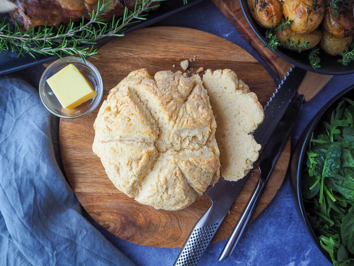 Damper 1 Damper on a chopping board with butter, bread knife and dishes on the sides, beetroot salad, mini roasted potatoes and slow cooker lam shoulder.