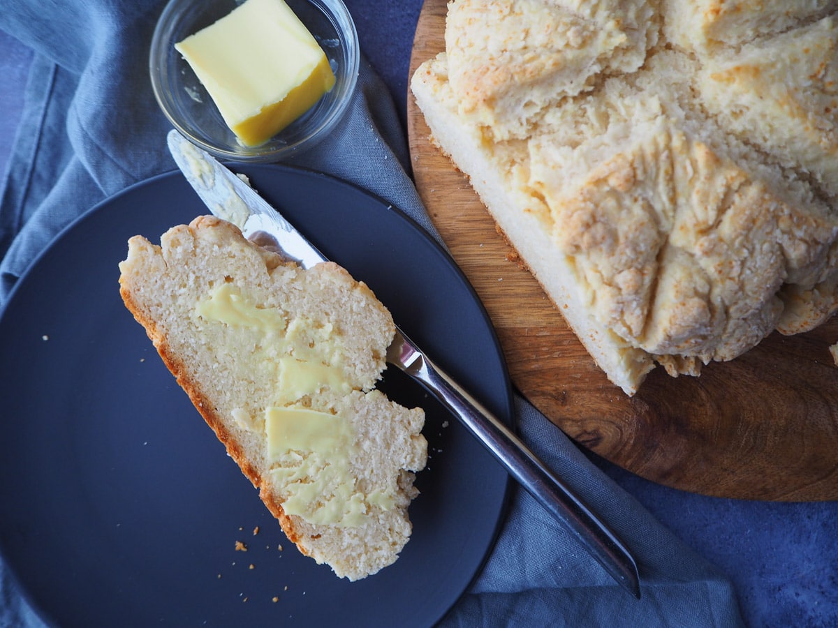 Damper 12 Cut slice of buttered damper on a plate with butter knife, next rest of damper on a chopping board, with butter in dish.