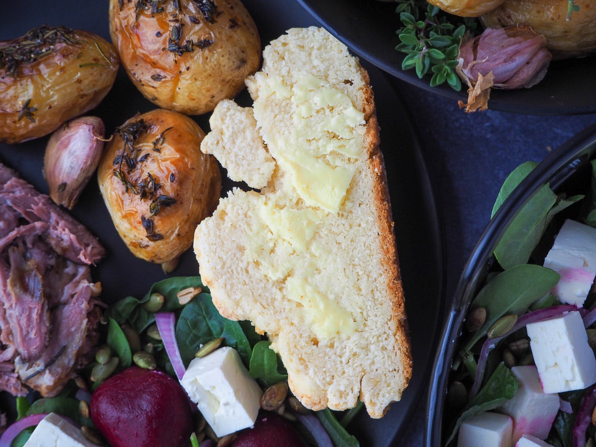 Damper 13 Slice of buttered damper on a plate with beetroot salad, mini roasted potatoes, slow cooker lamb shoulder and larger dishes on the side.