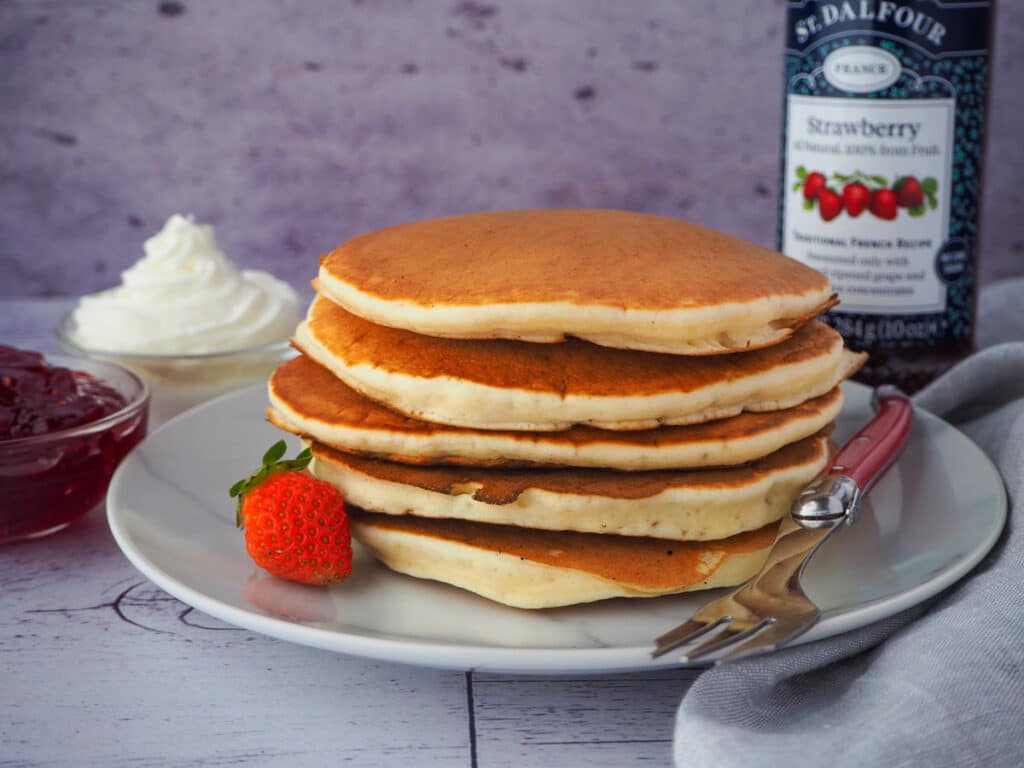 Pikelets 15 stack of pikelets on a plate, with a fresh strawberry and for, and whipped cream and jar of strawberry jam in the background,