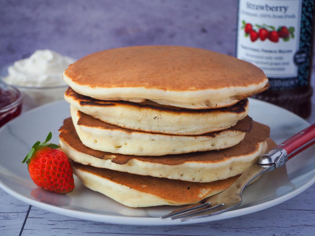 Pikelets 1 stack of pikelets on a plate, with a fresh strawberry and for, and whipped cream and jar of strawberry jam in the background,