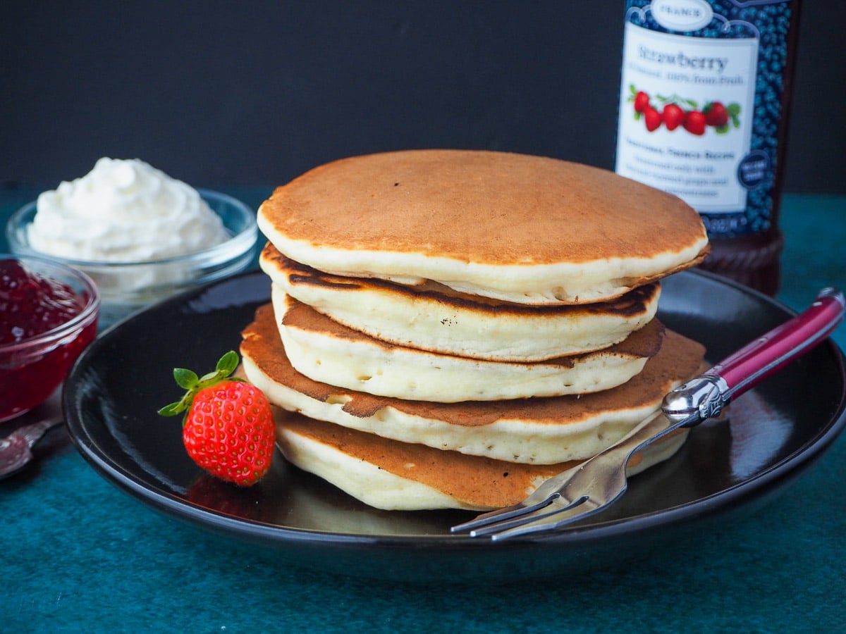 Pikelets 2 stack of pikelets on a plate, with a fresh strawberry and for, and whipped cream and jar of strawberry jam in the background,
