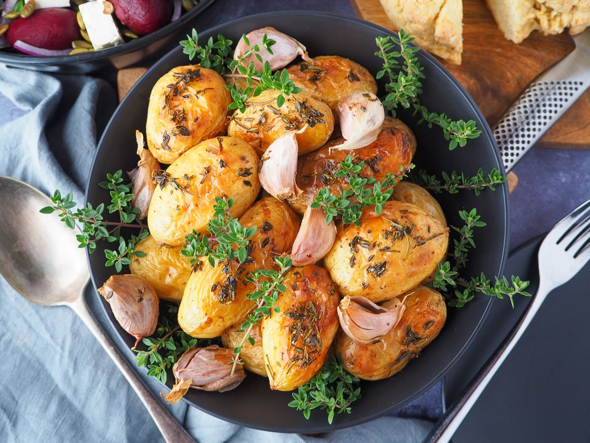 Roasted mini potatoes 13 Roasted mini potatoes in a serving dish garnished with fresh herbs, silver serving spoon to the side, fresh damper, pat of butter, beetroot salad and slow cooker roast lamb on the side.