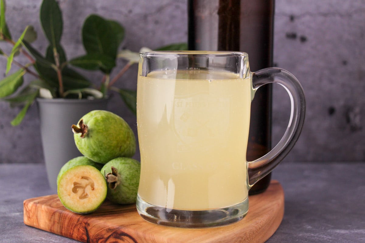 Feijoa fizz 11 Tankard glass of feijoa fizz on a board, with bottle of feijoa fizz, fresh feijoas and feijoa leaves.