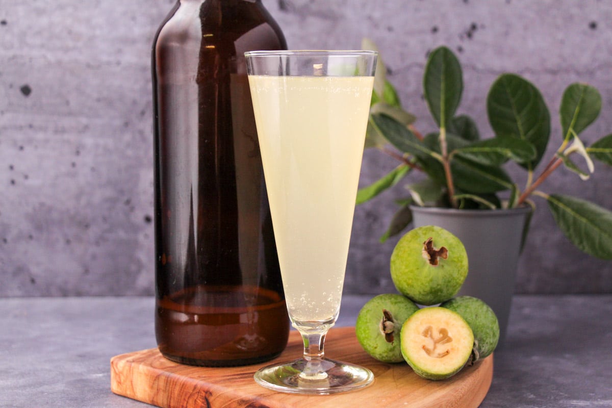 Feijoa fizz 12 Tall glass of feijoa fizz on a board, with bottle of feijoa fizz, fresh feijoas and feijoa leaves.