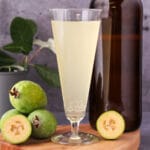 Feijoa fizz 13 Close up tall glass of feijoa fizz on a board, with bottle of feijoa fizz, fresh feijoas and feijoa leaves.