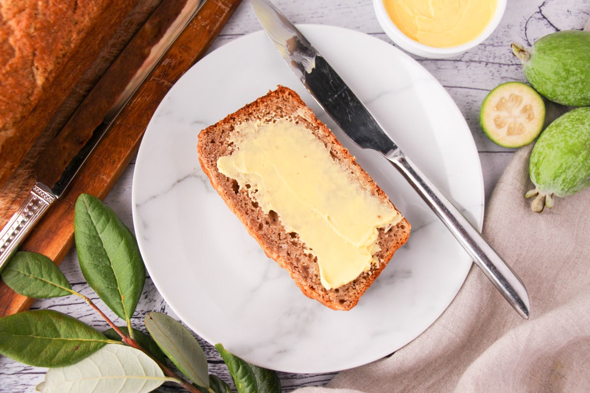 Slice of buttered feijoa loaf on a plate with butter knife, fresh feijoas and leaves and a tea towel.