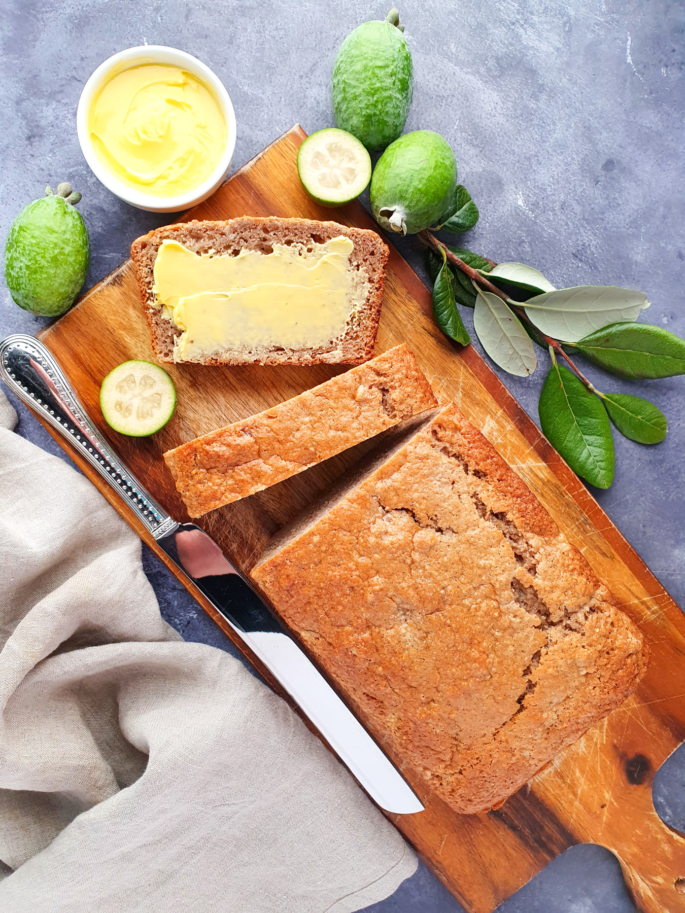 Sliced feijoa loaf on a chopping board, with one buttered slice, fresh feijoas and leaves and a butter knife.