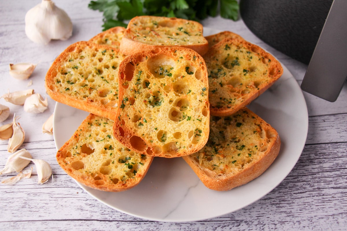 Air Fryer Garlic Bread 1 Stack of air fryer garlic bread on a plate, with air fryer basket and fresh garlic cloves to the sides.