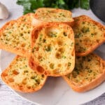 Air Fryer Garlic Bread 12 Close up of stack of air fryer garlic bread on a plate, with air fryer basket and fresh garlic cloves to the sides.