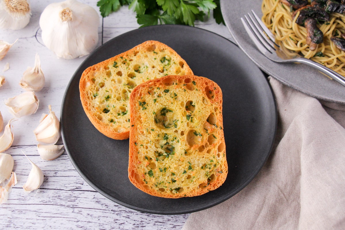 Air Fryer Garlic Bread 2 Air fryer garlic bread on a plate, with fresh garlic cloves, fresh parsley and a plate of pasta on the sides.