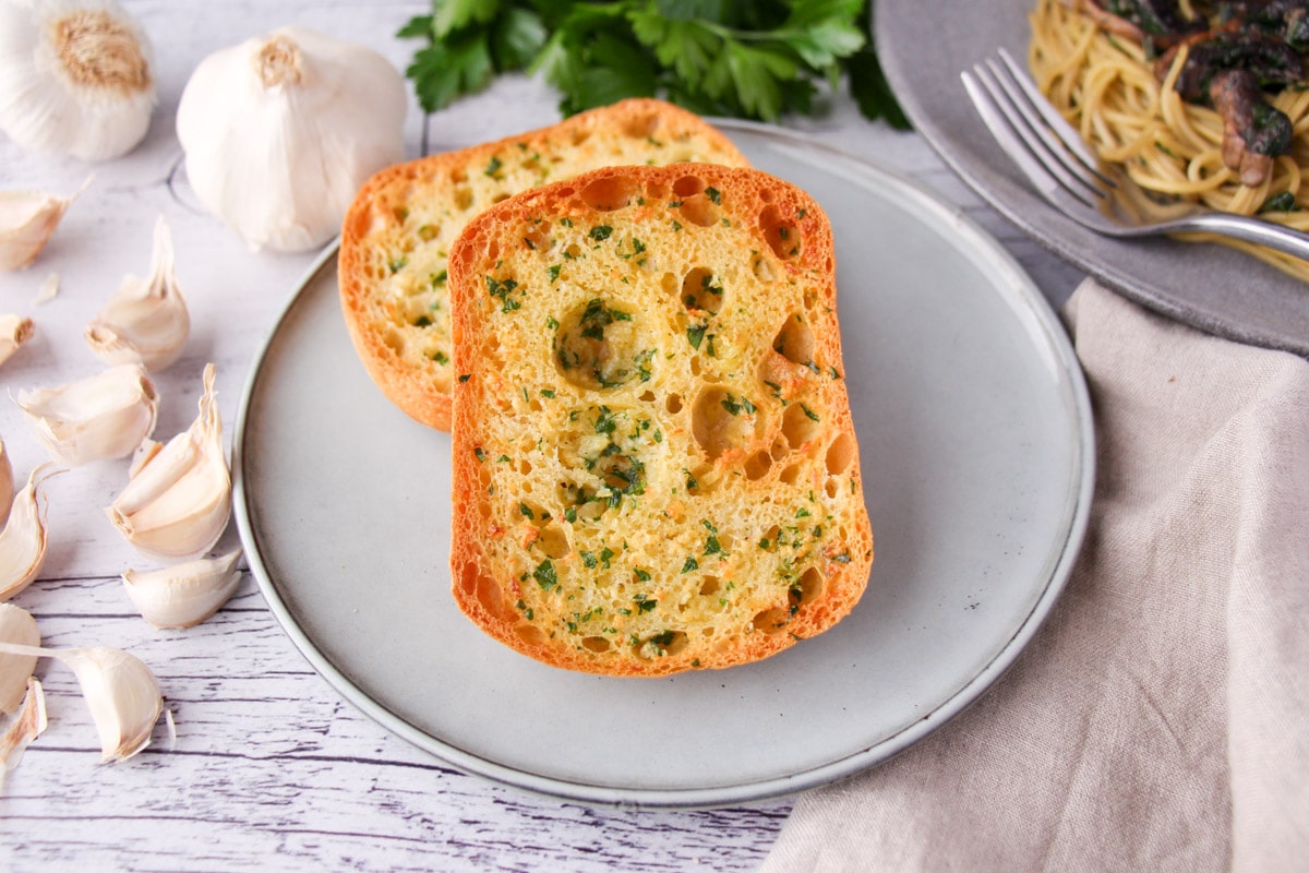 Air Fryer Garlic Bread 10 Air fryer garlic bread on a plate, with fresh garlic cloves, fresh parsley and a plate of pasta on the sides.
