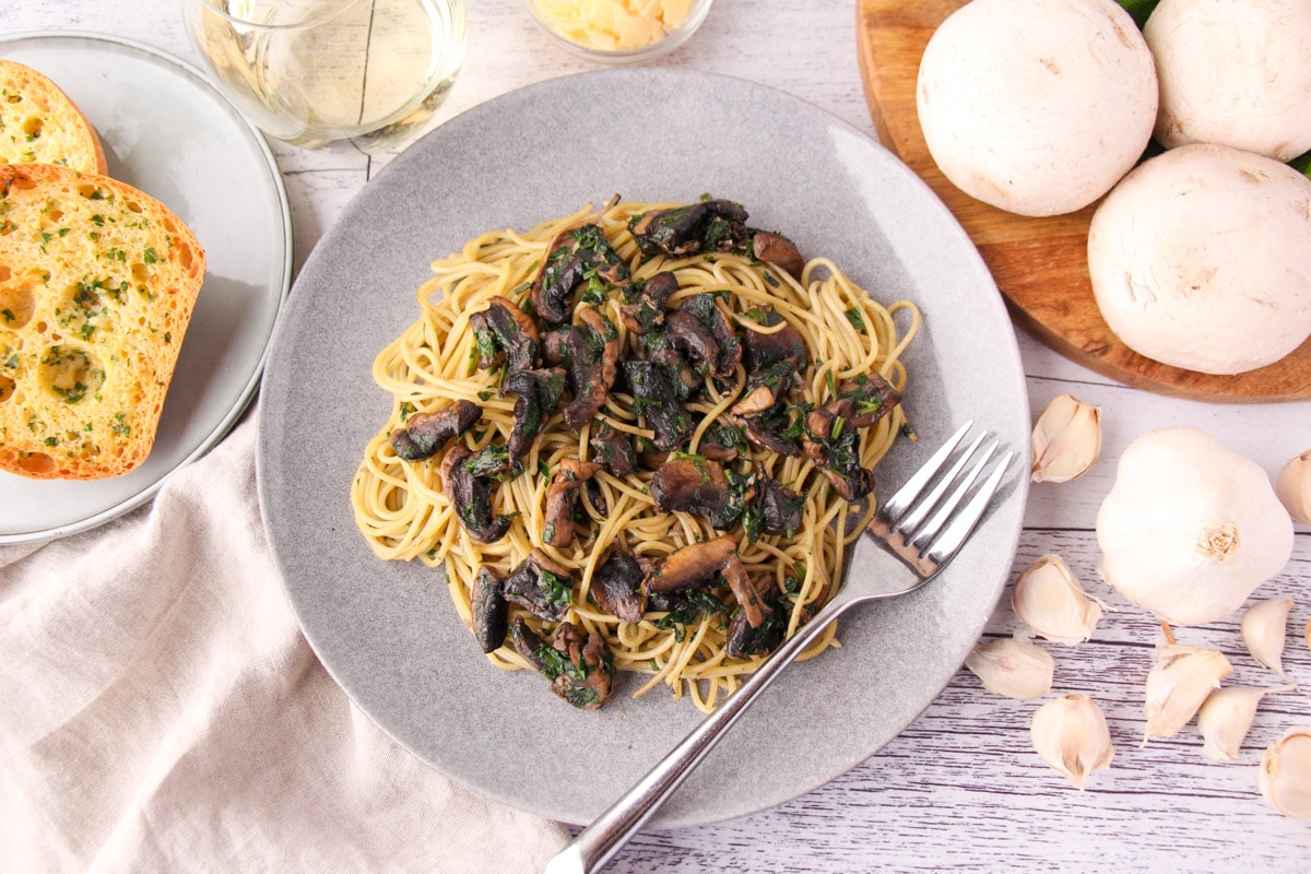 Mushroom Aglio Olio 16 Mushroom aglio olio on a plate with a fork, with garlic bread, fresh garlic, mushrooms and parsley, a glass of white wine and shaved parmesan around the edges.