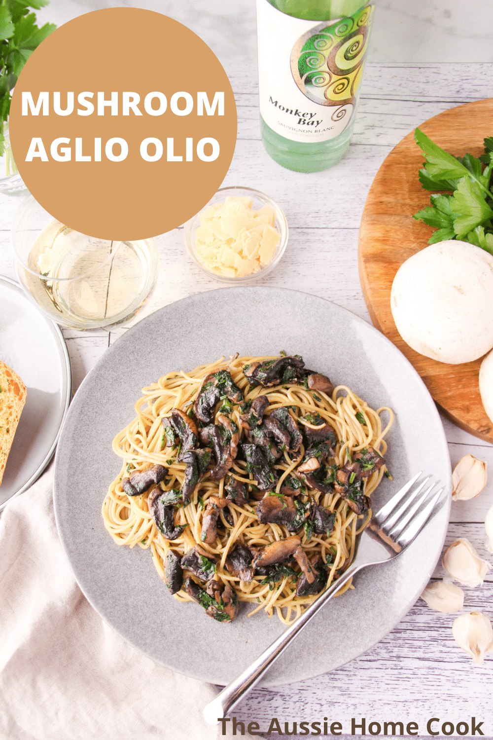 Mushroom Aglio Olio 18 Mushroom aglio olio on a plate with a fork, with garlic bread, fresh garlic, mushrooms and parsley, a glass and bottle of white wine and shaved parmesan around the edges., with text overlay, mushroom aglio olio, The Aussie Home Cook.