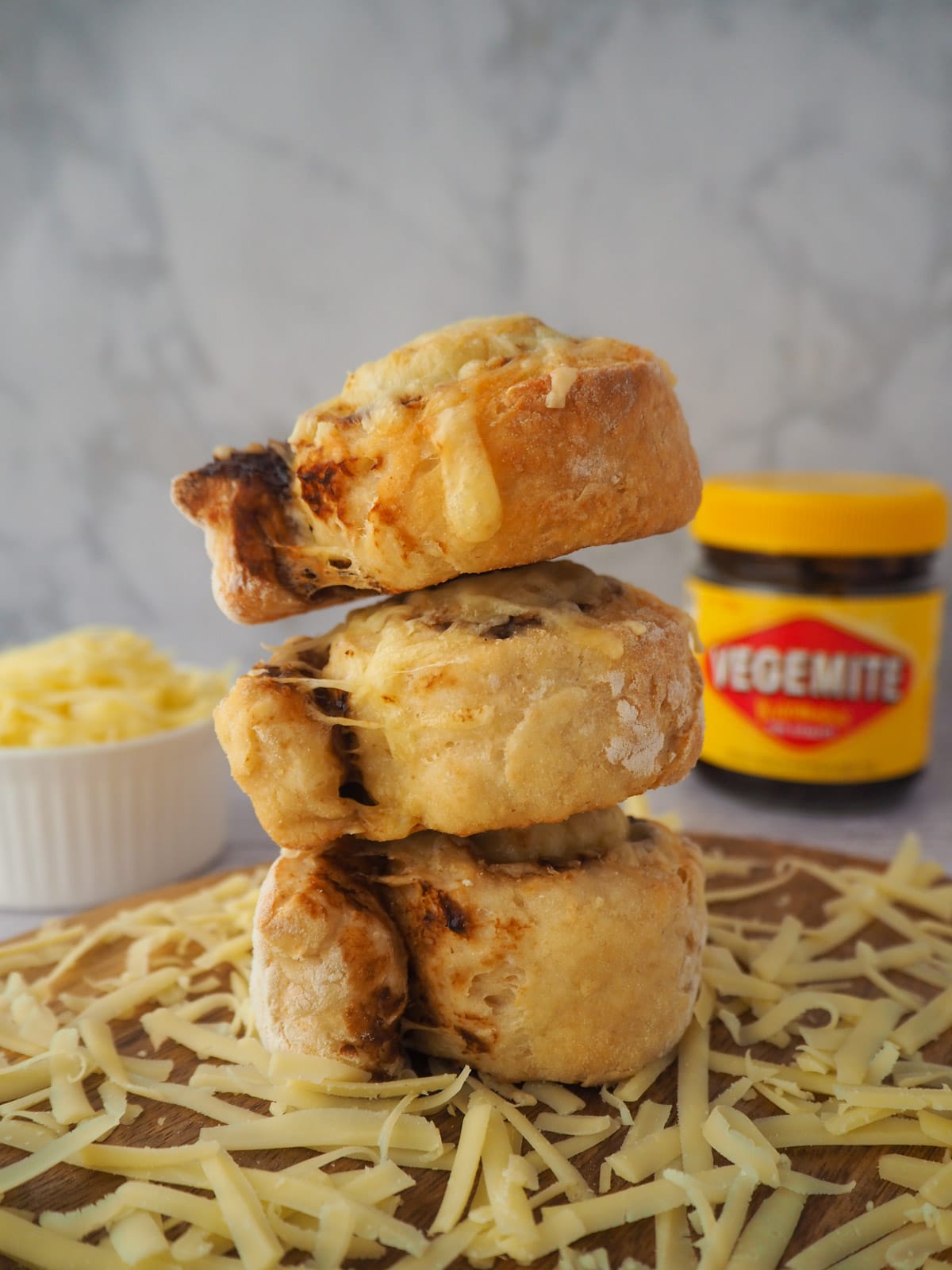 Cheesymite Scrolls 18 Stack of scrolls surrounded by grated cheese, with bowl of grated cheese and jar of vegemite in the background.
