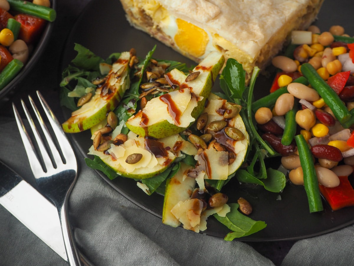Rocket and Pear Salad 11 Serving of salad on a plate with bacon and egg pie and four bean salad, knife and fork to the side and four bean salad in the background.