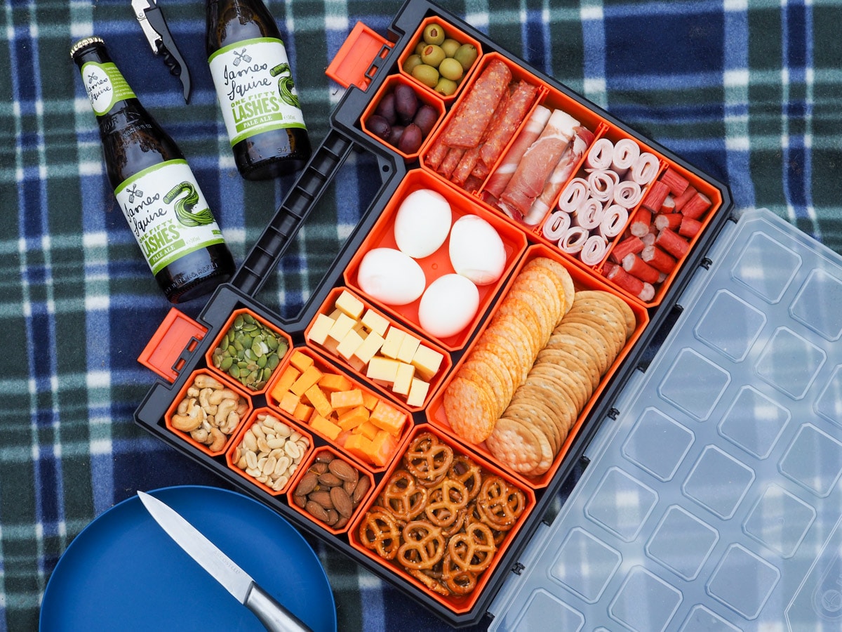 Picnic Charcuterie Board 1 Multi compartment container filled with various charcuterie food, on a picnic blanket with a plate and drinks.