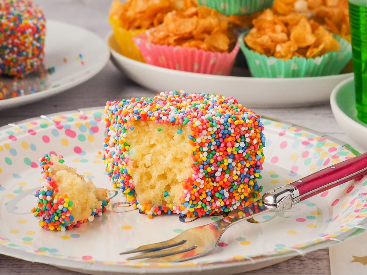 Fairy Bread Lamingtons 10 Lamington with one piece out on a plate with a fork and party food in the background.