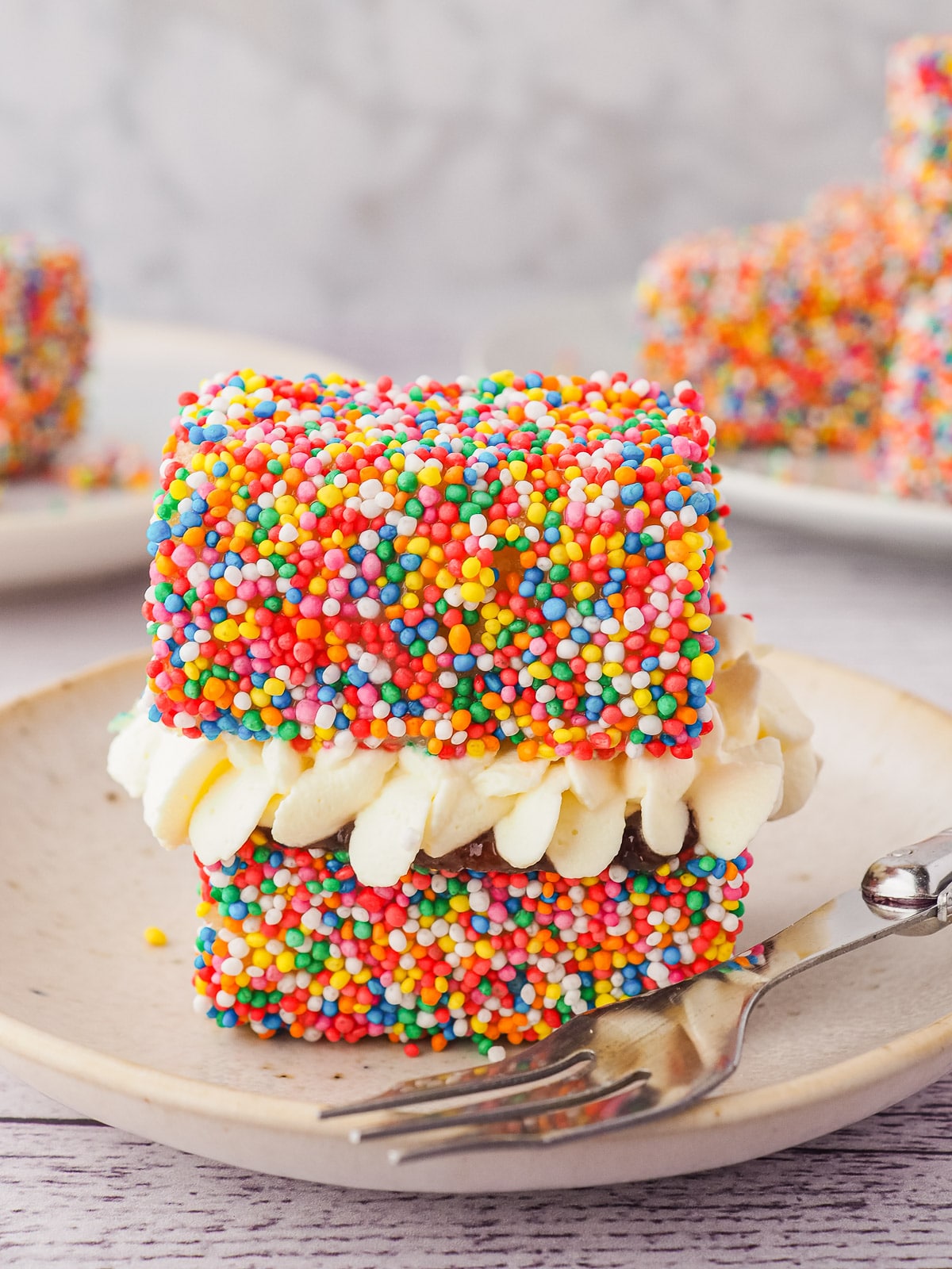 Fairy Bread Lamingtons 13 Lamington with jam and cream on a plate with a fork, and plate of lamingtons in the backgroun.
