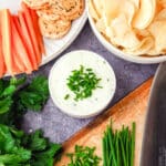 Sour Cream Dip 9 Sour cream dip garnished with fresh chives, with a bowl of chips, plate of crackers and carrot sticks, and chopping board with freshly chopped chives.