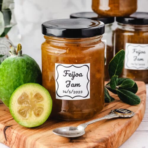 Feijoa Jam 30 Jar of feijoa jam on a serving board with fresh feijoas and leaves on the side, more jars and a plate of toasted muffins in the background.