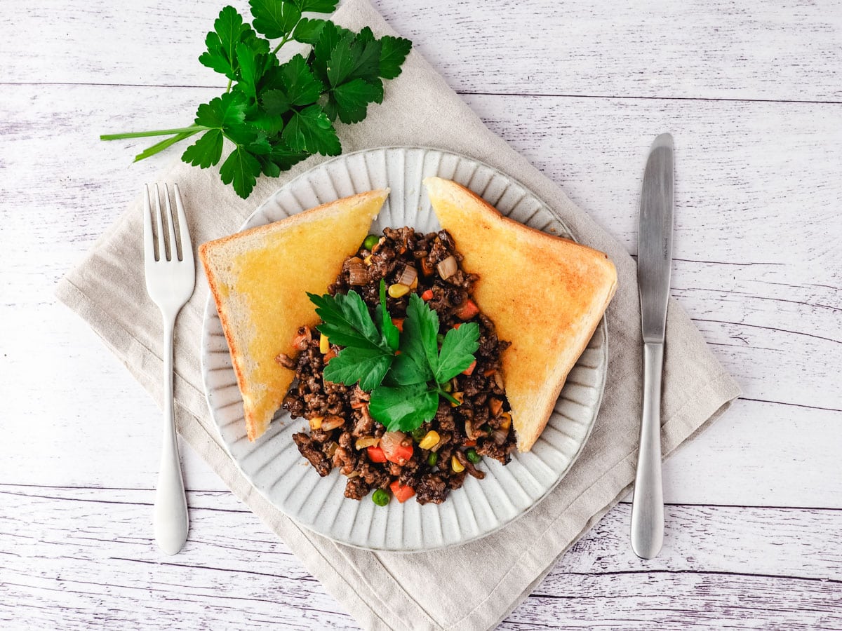 Savory Mince 6 Savory mince on a plate with buttered toast on the side, garnished with flat parsley, with a knife and fork on the side.