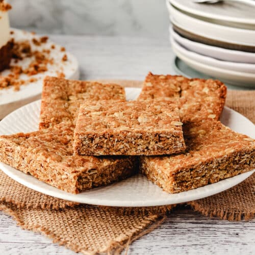 Anzac Slice 26 A pile of pieces of Anzac slice on a plate, with a pile of plates in the background.