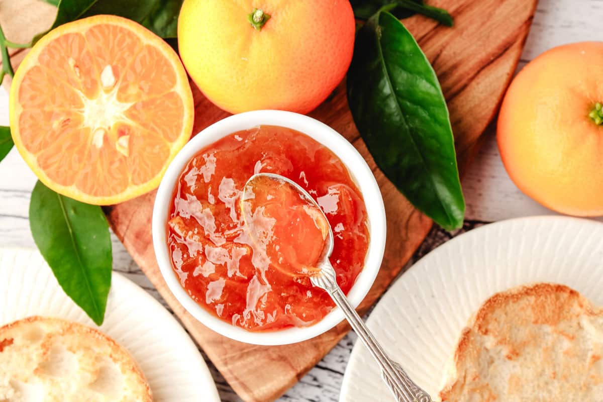 Mandarin Marmalade 6 Top down view of mandarin marmalade in a bowl with a spoon, with toasted English muffins and fresh mandarins on the side.